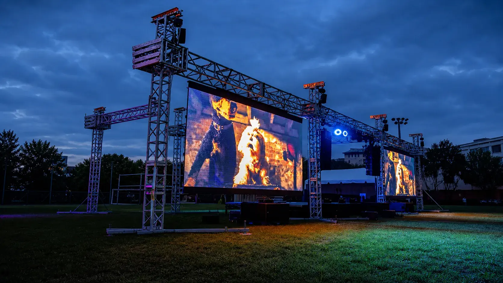 Outdoor LED screen illuminated at dusk beside event truss