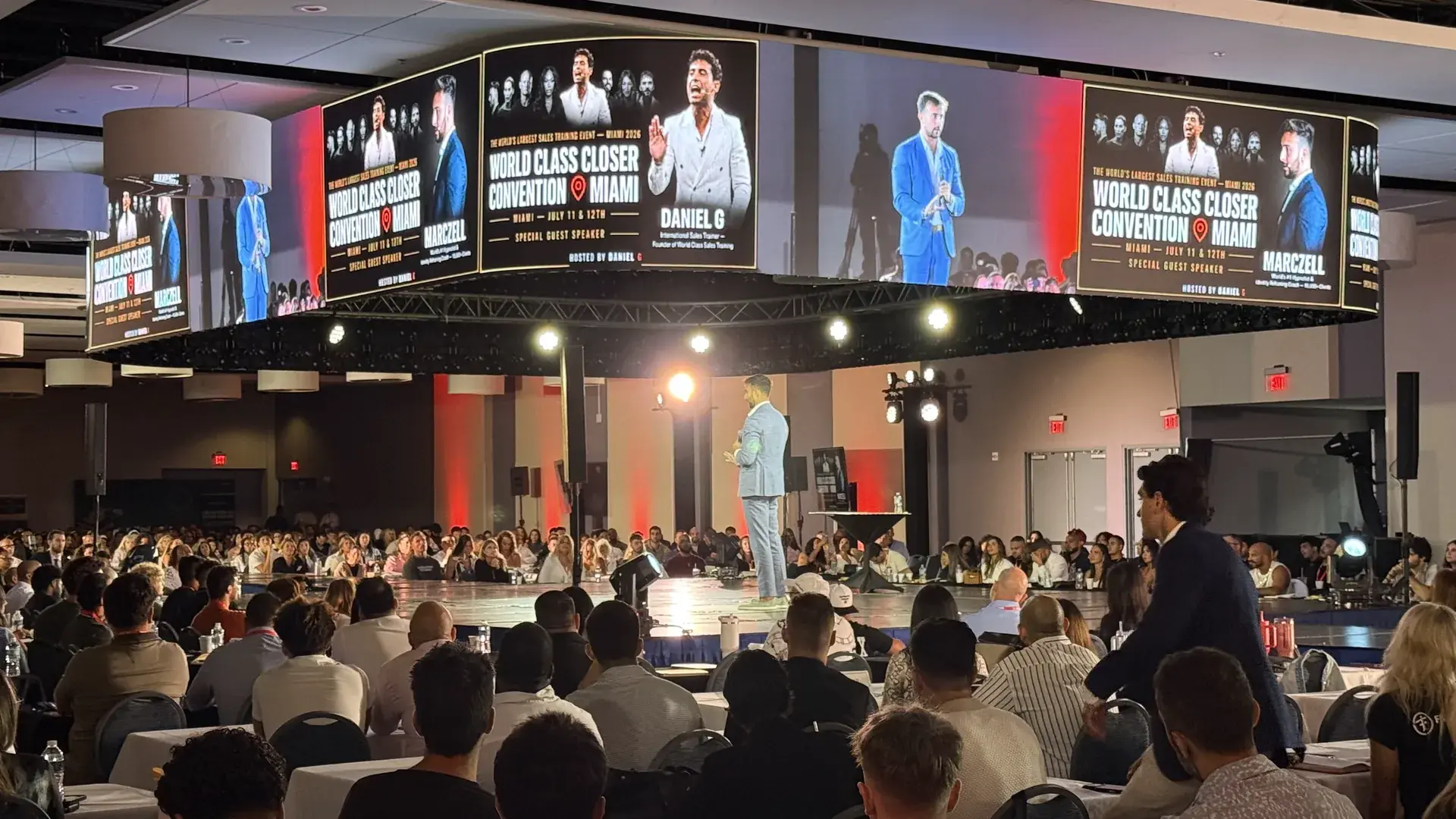 Conferencista en tarima central rodeado de pantallas LED suspendidas y una audiencia completa