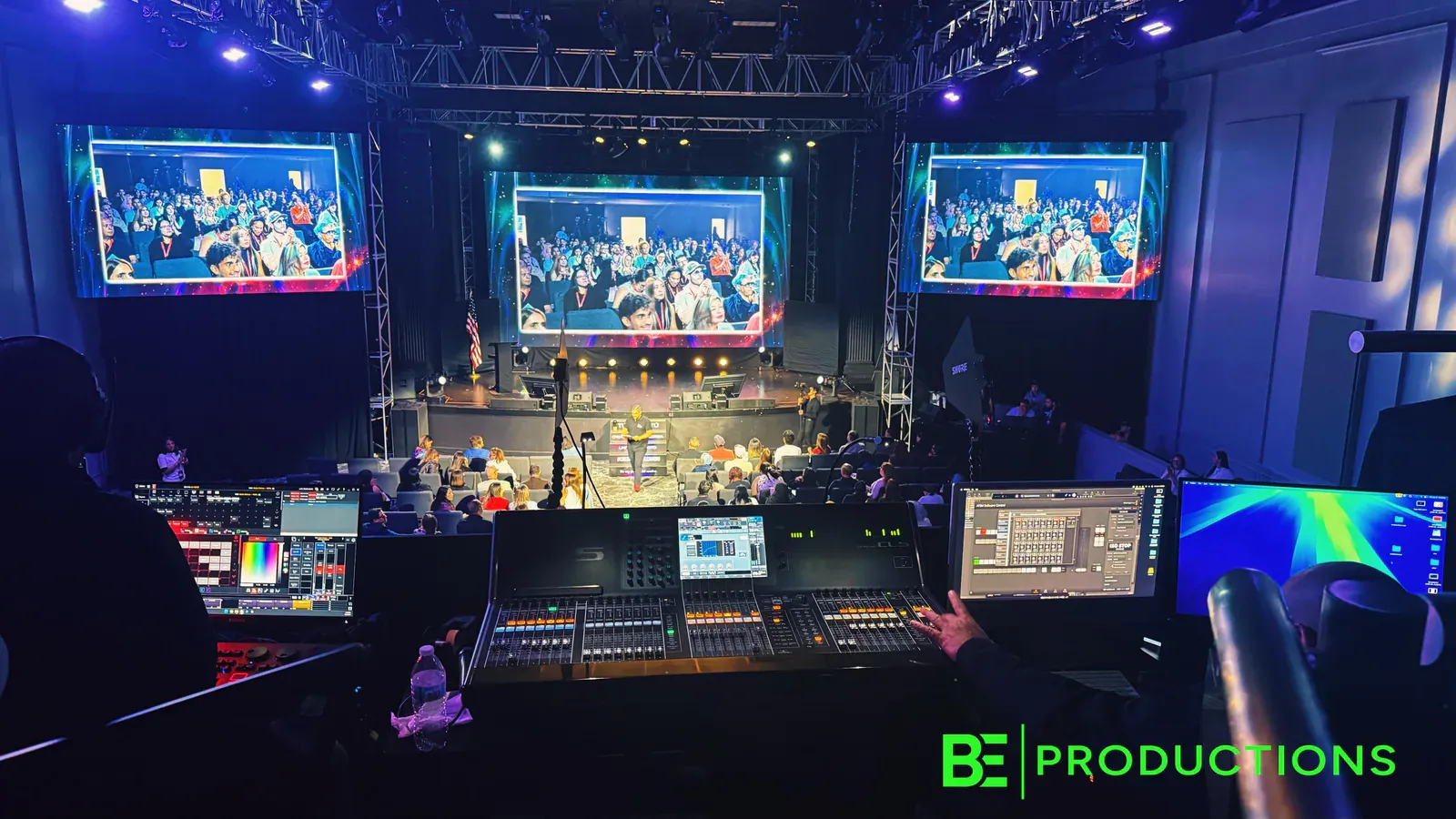Live production controls facing a conference audience and three LED screens